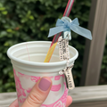 White cup with pink design, straw with blue bow, and motivational charm held by a hand.